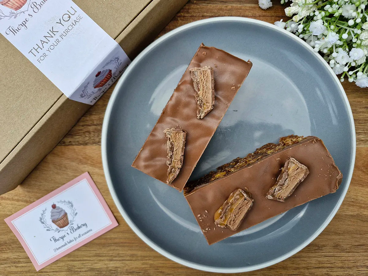 Two Mars Bar crispy slices topped with mars bar pieces on a plate beside a Thorpe’s Bakery treat box.