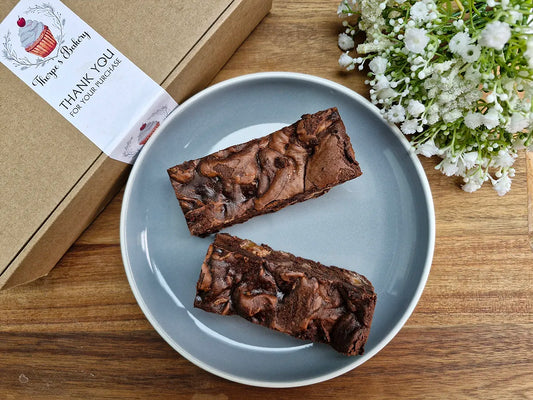 Two large rich brownie slices on a plate beside a Thorpe’s Bakery treat box on a rustic wooden surface.