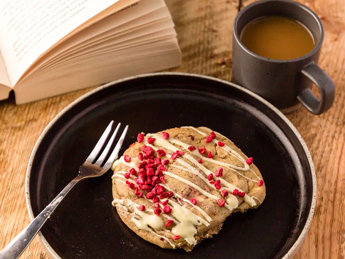 Large white chocolate and raspberry cookie on a plate beside a cup of tea and an open book. 