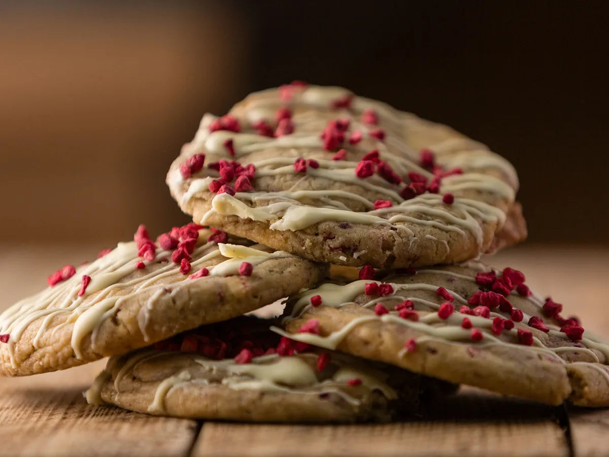 Stack of cookies drizzled with white chocolate and topped with raspberry pieces on a rustic wooden surface.