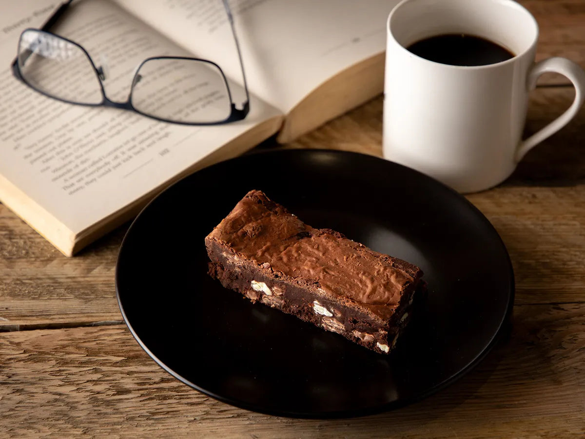 Chocolate brownie slice on a plate beside an open book and cup of coffee.